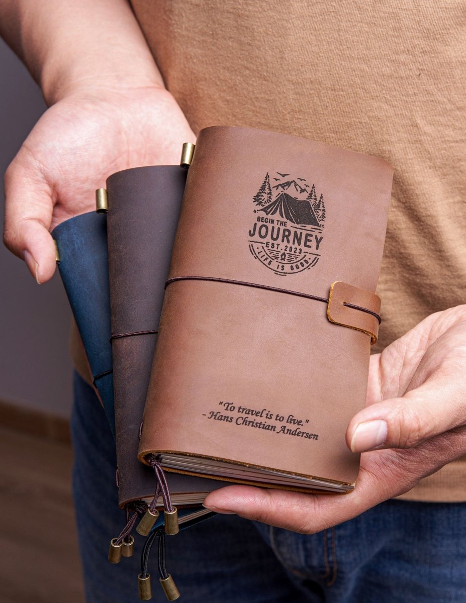 Person holding a handcrafted leather travel journal with a mountain adventure engraving and quote, showcasing the pocket-size design and elastic closure for travel journaling.
