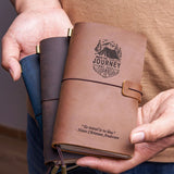 Person holding a handcrafted leather travel journal with a mountain adventure engraving and quote, showcasing the pocket-size design and elastic closure for travel journaling.