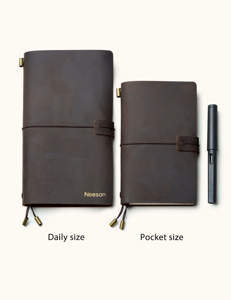 Two handmade coffee brown leather travel notebooks in daily and pocket sizes with elastic closure and custom name engraving, shown with a pen for size reference.