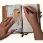 Person writing in a handmade leather travel journal with a grid refill while using the notebook for collage scrapbooking and journaling.