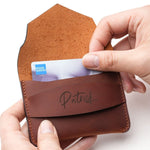 Person inserting a credit card into a handcrafted brown leather wallet with personalized name engraving, showing the slim minimalist card holder in use.