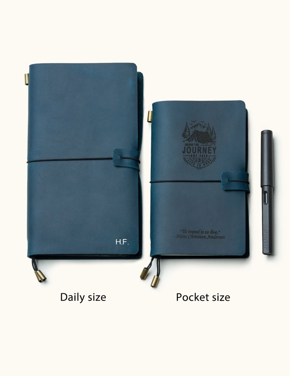 Two handmade navy blue leather travel notebooks in daily and pocket sizes with elastic closure, custom engraving, and refillable design, shown with pen for size reference.