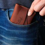 Handcrafted brown leather wallet with personalized engraving being placed into a jeans pocket, showing slim minimalist card holder design for easy everyday carry.