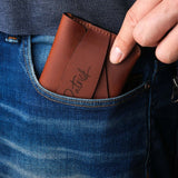 Handcrafted brown leather wallet with personalized engraving being placed into a jeans pocket, showing slim minimalist card holder design for easy everyday carry.
