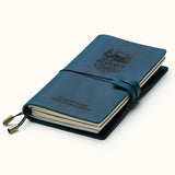 Handcrafted navy blue leather travel journal with elastic closure, custom engraved mountain adventure design, refillable pages, and brass charms, perfect for travel notes and journaling.
