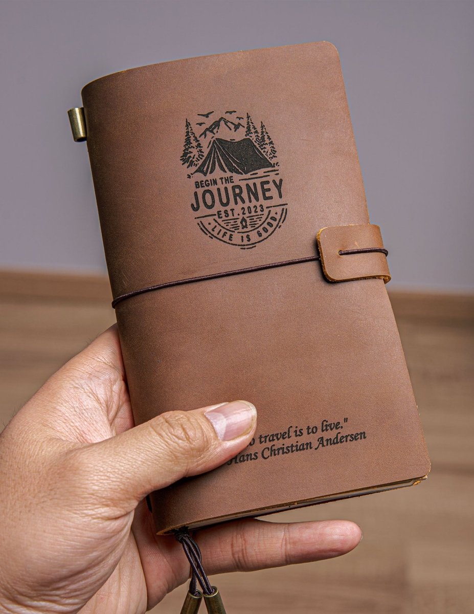 Hand holding a handcrafted vintage brown leather travel journal with elastic closure, custom engraved mountain adventure design and inspirational quote, showing pocket-size travel notebook for journaling.