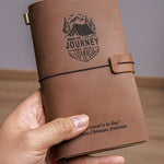 Hand holding a handcrafted vintage brown leather travel journal with elastic closure, custom engraved mountain adventure design and inspirational quote, showing pocket-size travel notebook for journaling.