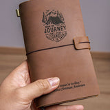 Hand holding a handcrafted vintage brown leather travel journal with elastic closure, custom engraved mountain adventure design and inspirational quote, showing pocket-size travel notebook for journaling.