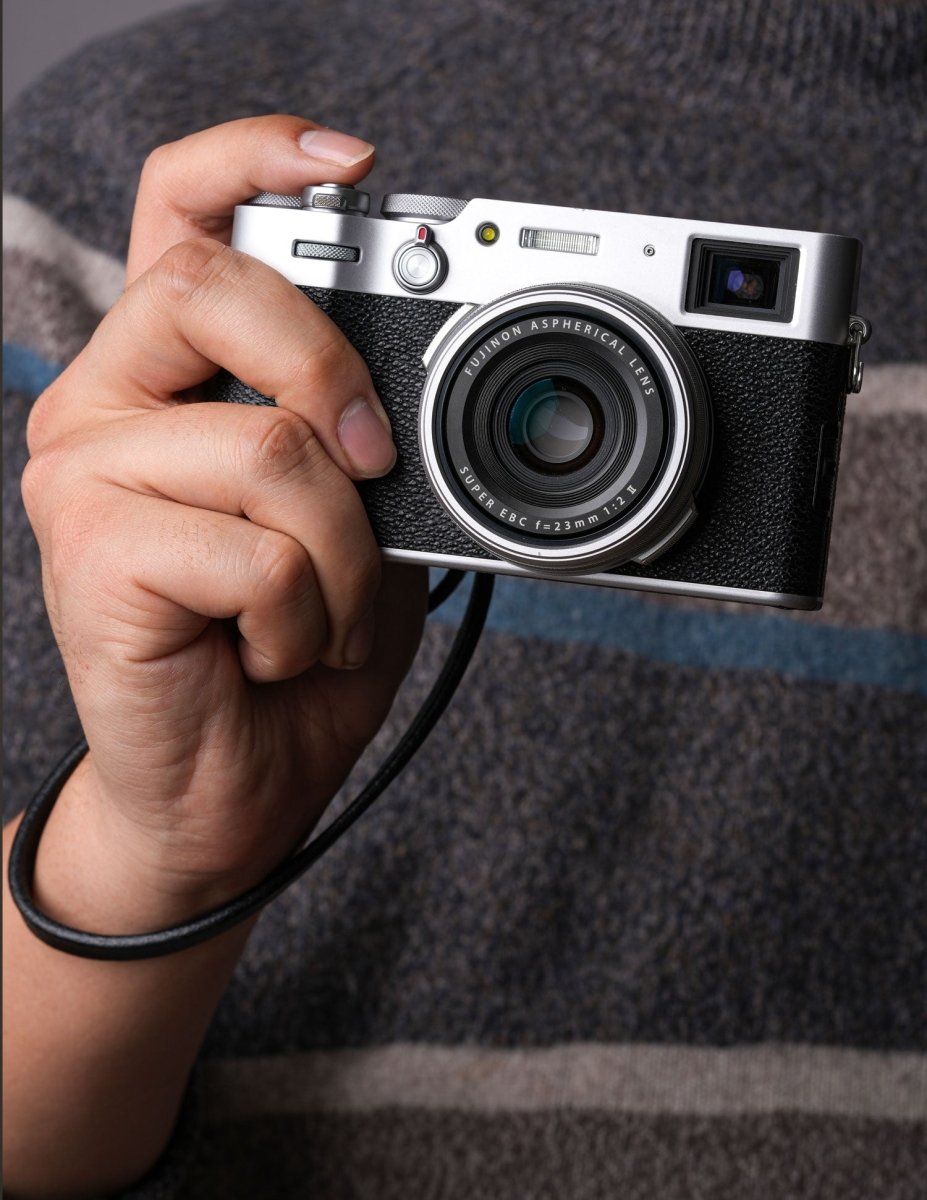 Close-up of a photographer holding a Fujifilm X100 series camera with a black leather wrist strap looped around the wrist, ready to shoot