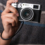 Close-up of a photographer holding a Fujifilm X100 series camera with a black leather wrist strap looped around the wrist, ready to shoot