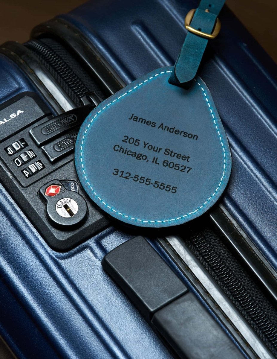 Blue leather luggage tag with engraved name and address shown attached to a suitcase