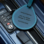 Blue leather luggage tag with engraved name and address shown attached to a suitcase