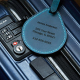 Blue leather luggage tag with engraved name and address shown attached to a suitcase