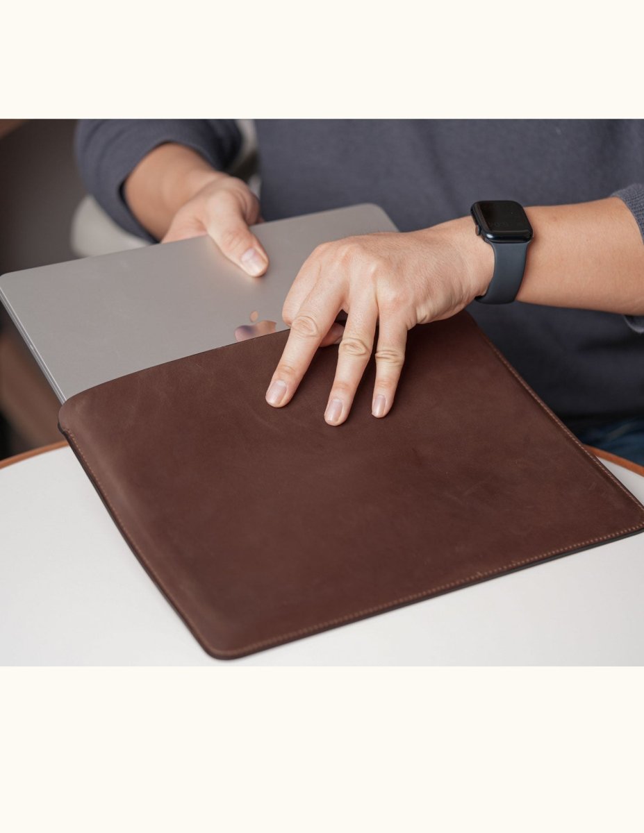 Person inserting a MacBook into a customizable coffee color full-grain leather sleeve, shown in real-life use