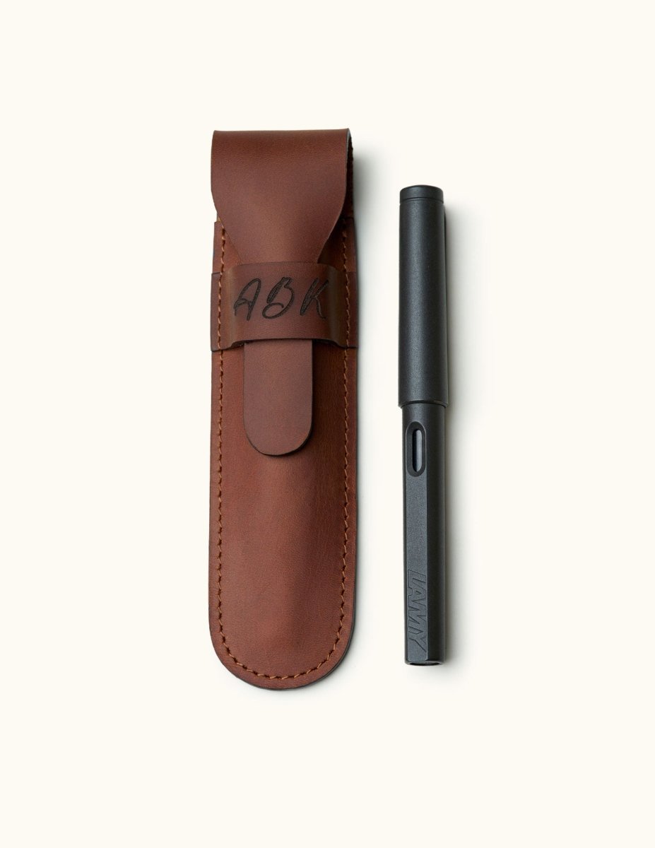 Personalized leather fountain pen case with engraved initials, shown next to a black LAMY pen. Crafted from full-grain leather with flap closure and fine stitching