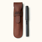 Personalized leather fountain pen case with engraved initials, shown next to a black LAMY pen. Crafted from full-grain leather with flap closure and fine stitching