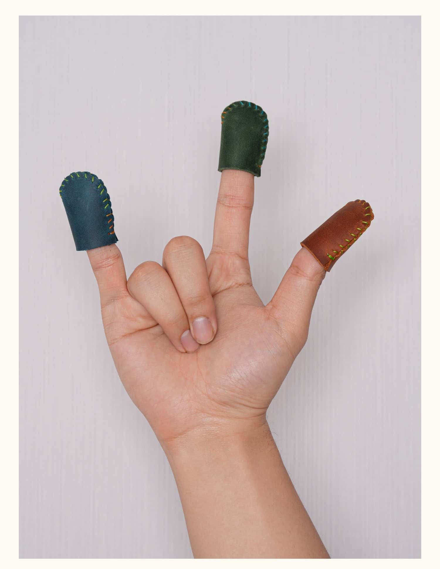 Hand showing three fingers wearing full-grain leather thimbles in blue, green, and brown, ideal for sewing or crafting.