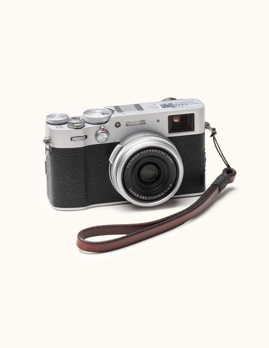 Angled view of a Fujifilm X100V camera with a handcrafted brown leather wrist strap attached, combining vintage design with everyday functionality