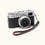 Angled view of a Fujifilm X100V camera with a handcrafted brown leather wrist strap attached, combining vintage design with everyday functionality