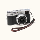 Angled view of a Fujifilm X100V camera with a handcrafted brown leather wrist strap attached, combining vintage design with everyday functionality
