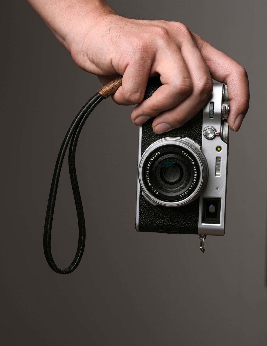 Photographer holding a Fujifilm camera by a black leather wrist strap with brown wax cord, demonstrating secure and stylish handheld use