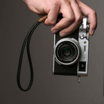 Photographer holding a Fujifilm camera by a black leather wrist strap with brown wax cord, demonstrating secure and stylish handheld use