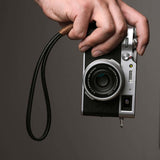 Photographer holding a Fujifilm camera by a black leather wrist strap with brown wax cord, demonstrating secure and stylish handheld use
