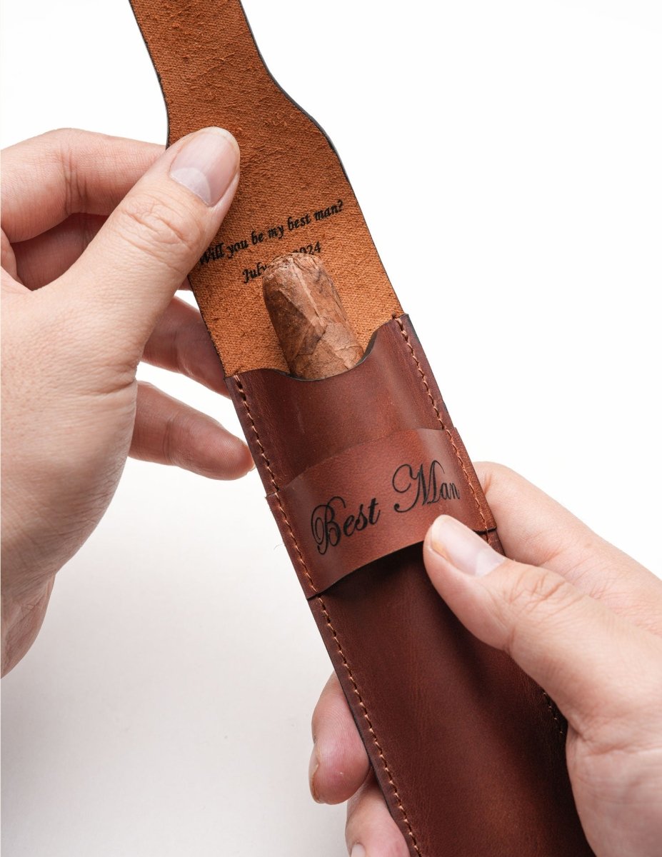 Hands placing a cigar into a personalized leather case engraved with "Best Man" on the outside and "Will you be my best man?" on the inner flap. A sentimental groomsman proposal gift idea