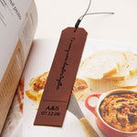 Brown personalized leather bookmark engraved with "To many more chapters together" and a custom date, placed inside an open cookbook next to slices of bread and a red dish
