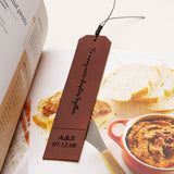 Brown personalized leather bookmark engraved with "To many more chapters together" and a custom date, placed inside an open cookbook next to slices of bread and a red dish