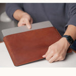 User placing a MacBook Pro into a vintage brown full-grain leather sleeve, shown in real-life usage scene