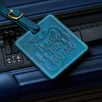 Handmade navy blue leather luggage tag with white stitching and brass buckle attached to a dark blue suitcase, embossed with “travel the world” design for travel identification.