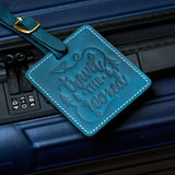 Handmade navy blue leather luggage tag with white stitching and brass buckle attached to a dark blue suitcase, embossed with “travel the world” design for travel identification.