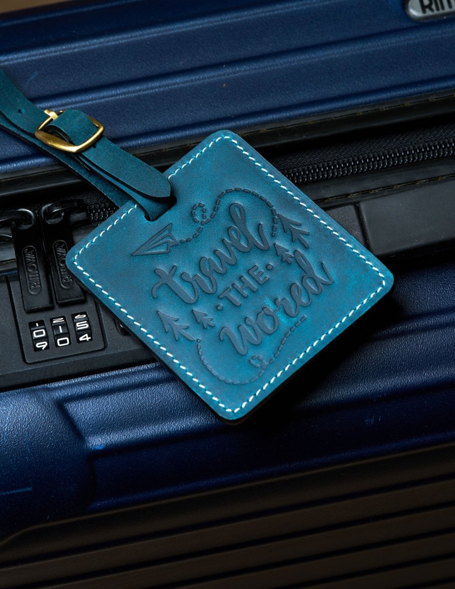 Handmade navy blue leather luggage tag with white stitching and brass buckle attached to a dark blue suitcase, embossed with “travel the world” design for travel identification.