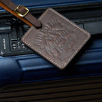 Handmade coffee brown leather luggage tag with white stitching and brass buckle attached to a dark blue suitcase, embossed with “And So The Adventure Begins” design for travel identification.