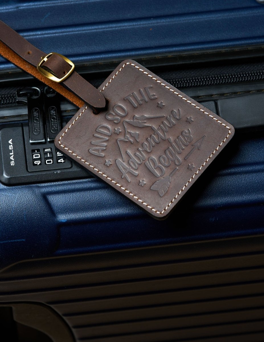 Handmade coffee brown leather luggage tag with white stitching and brass buckle attached to a dark blue suitcase, embossed with “And So The Adventure Begins” design for travel identification.