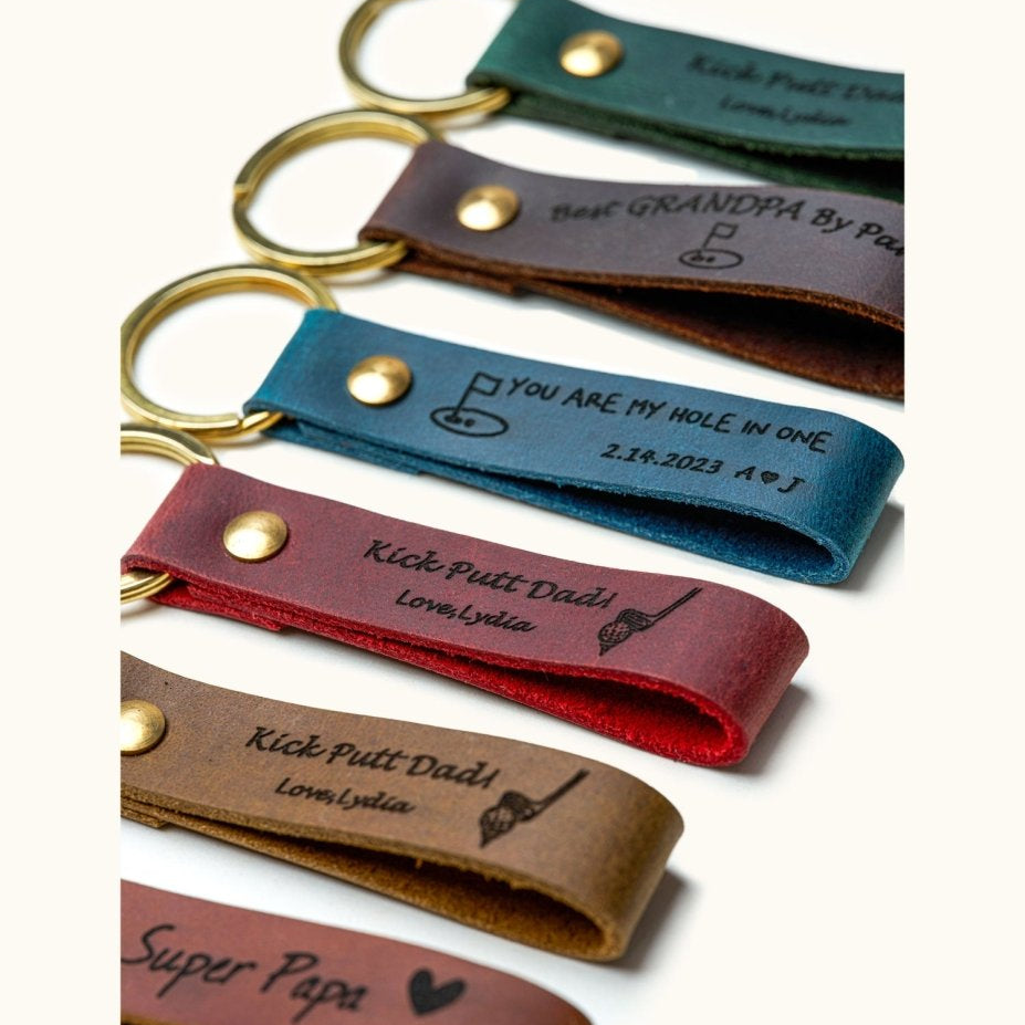 Close-up of handmade leather keychains with gold rings, engraved with golf-themed messages like “Kick Putt Dad” and “You Are My Hole In One,” perfect personalized gifts for golf dads, grandpas, and golf lovers.