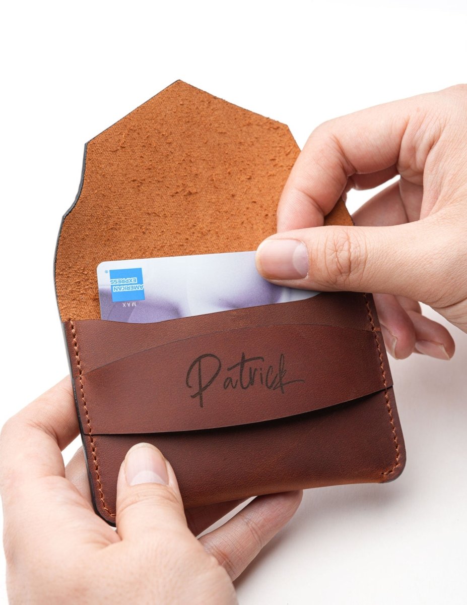 Person inserting a credit card into a handcrafted brown leather wallet with personalized name engraving, showing the slim minimalist card holder in use.