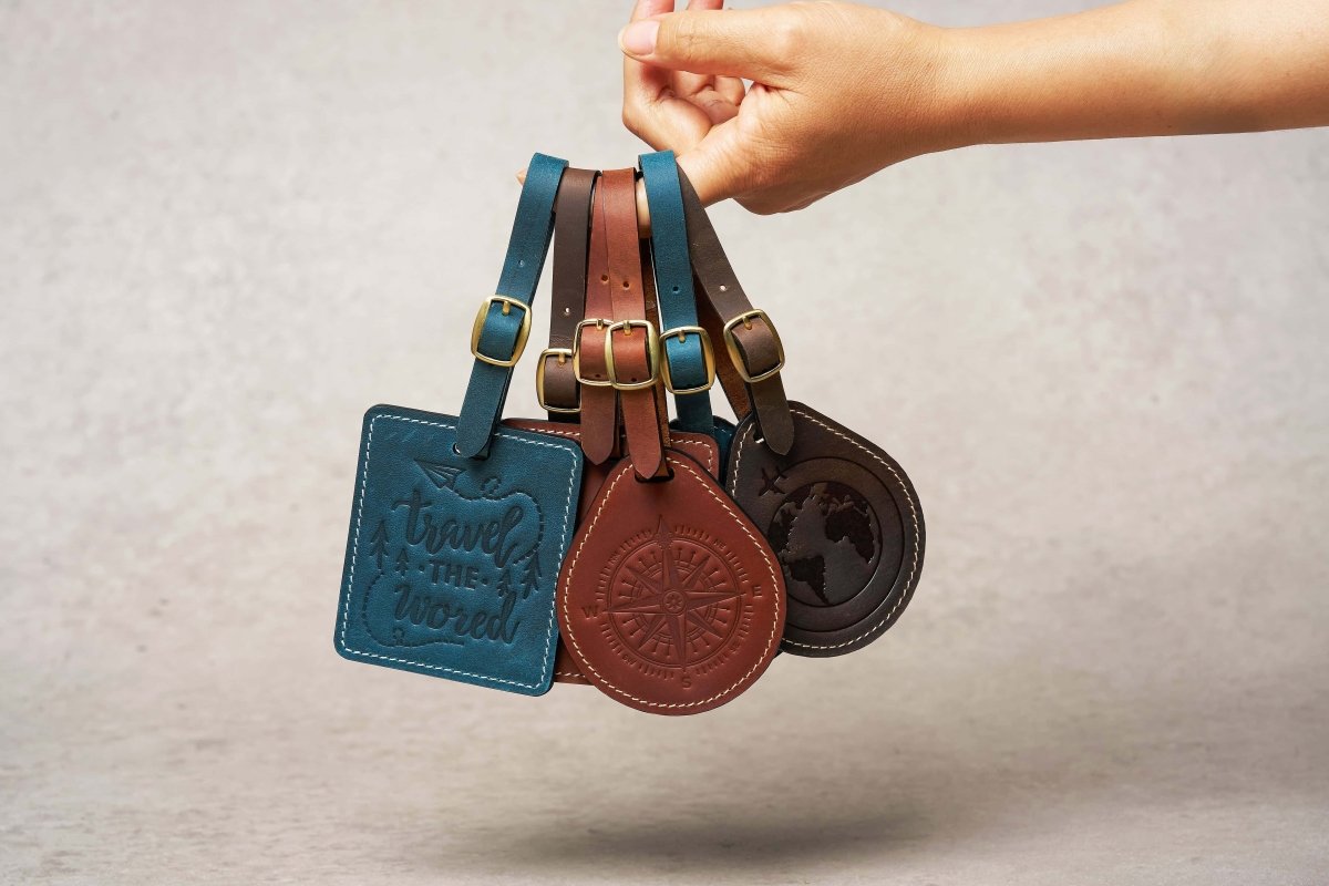Hand holding multiple handcrafted leather luggage tags in square and round shapes, in navy, brown, and coffee colors with embossed travel designs and brass buckles for travel identification.