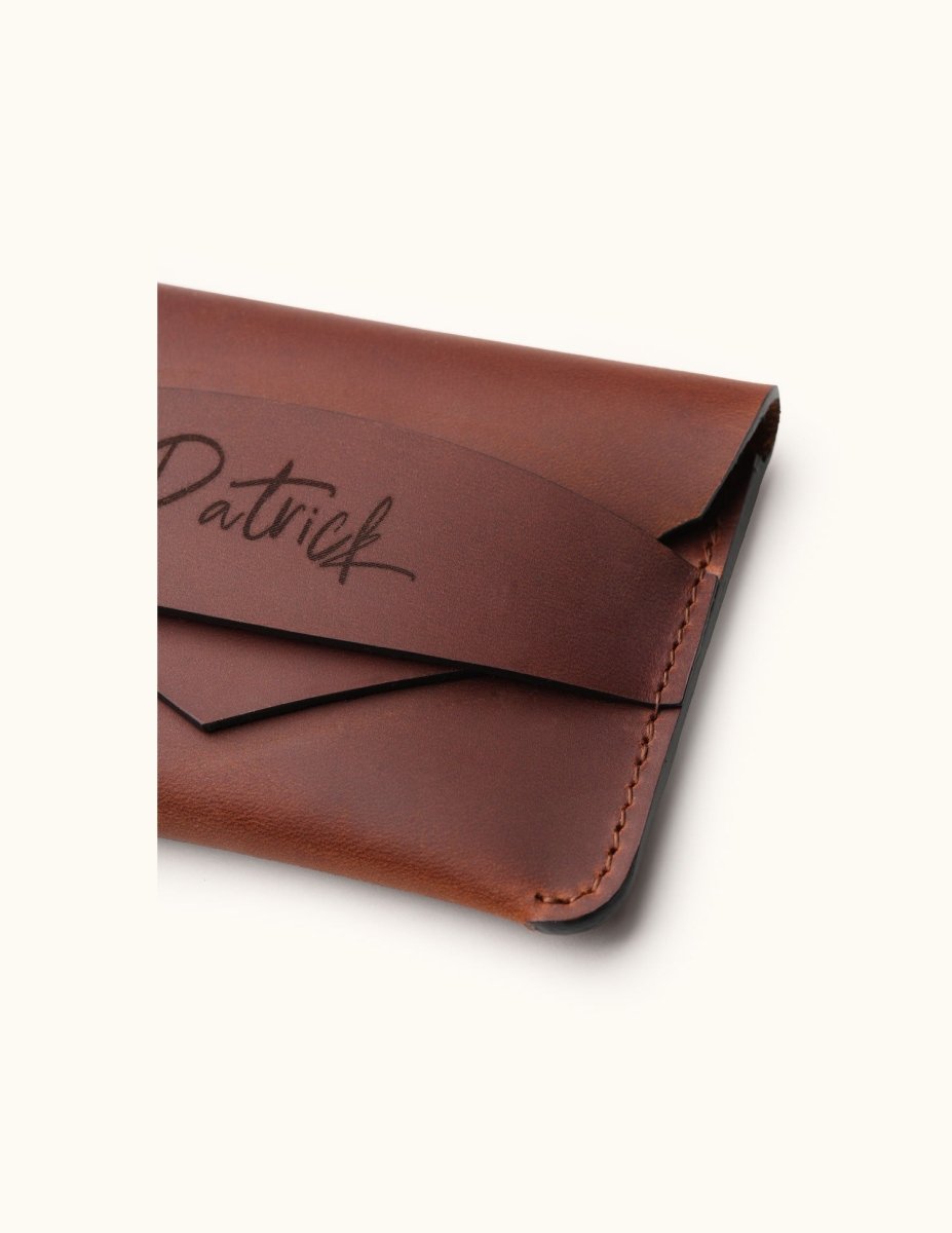 Close-up of a handcrafted brown leather wallet showing fine stitching, envelope flap design, and personalized name engraving on the slim minimalist card holder.