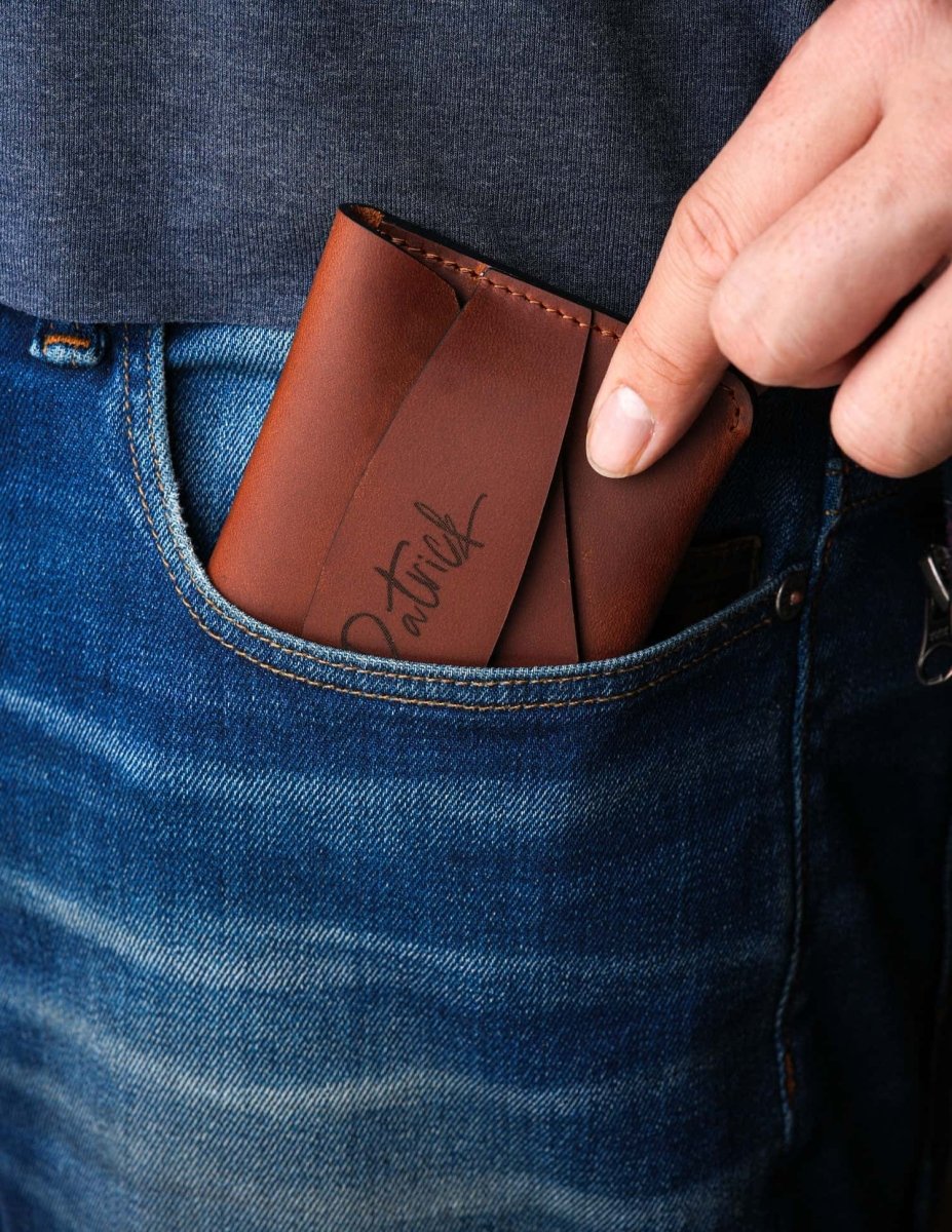 Handcrafted brown leather wallet with personalized engraving being placed into a jeans pocket, showing slim minimalist card holder design for easy everyday carry.