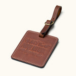 Handcrafted square brown leather luggage tag with white stitching, brass buckle strap, and embossed “let the adventure begin” quote for travel bag identification.