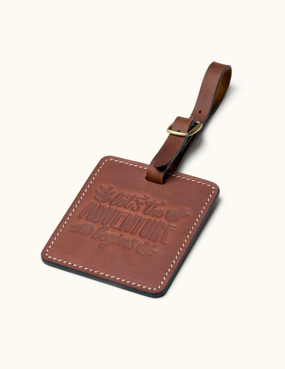 Handcrafted square brown leather luggage tag with white stitching, brass buckle strap, and embossed “let the adventure begin” quote for travel bag identification.