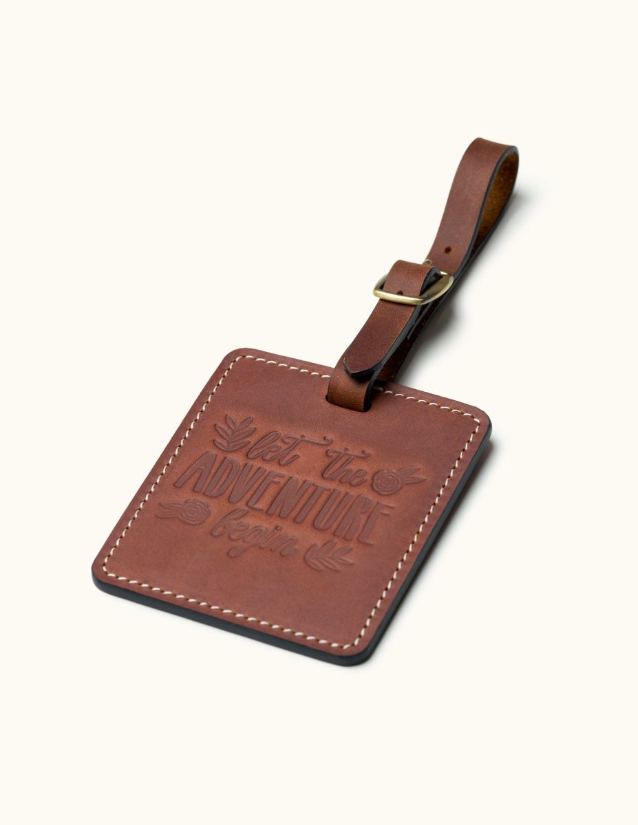 Handcrafted square brown leather luggage tag with white stitching, brass buckle strap, and embossed “let the adventure begin” quote for travel bag identification.