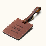 Handcrafted square brown leather luggage tag with white stitching and brass buckle strap, showing custom engraved personal contact information for travel bag identification.