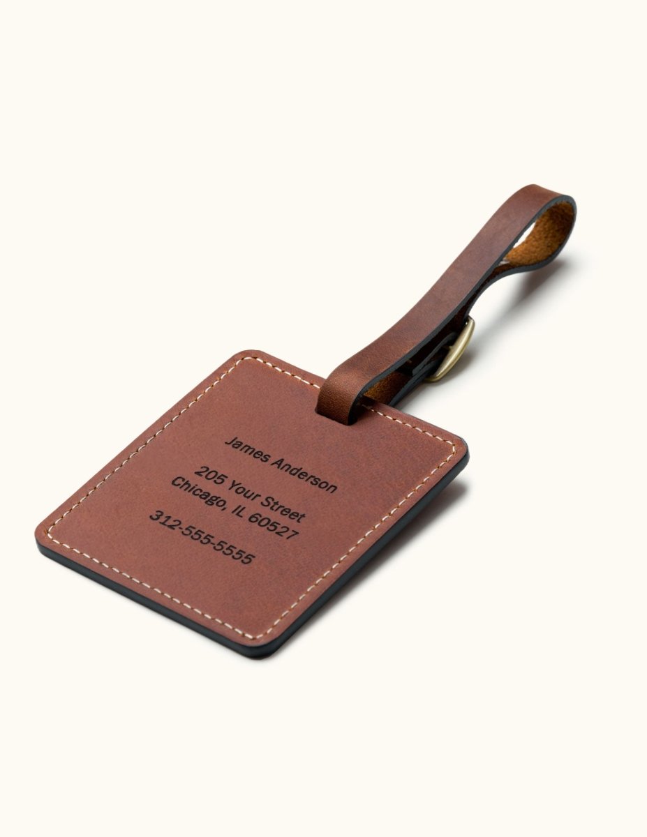 Handcrafted square brown leather luggage tag with white stitching and brass buckle strap, showing custom engraved personal contact information for travel bag identification.