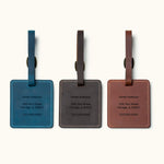 Three handcrafted square leather luggage tags in blue, coffee, and brown with custom engraved personal contact information, white stitching, and brass buckle straps for travel bag identification.