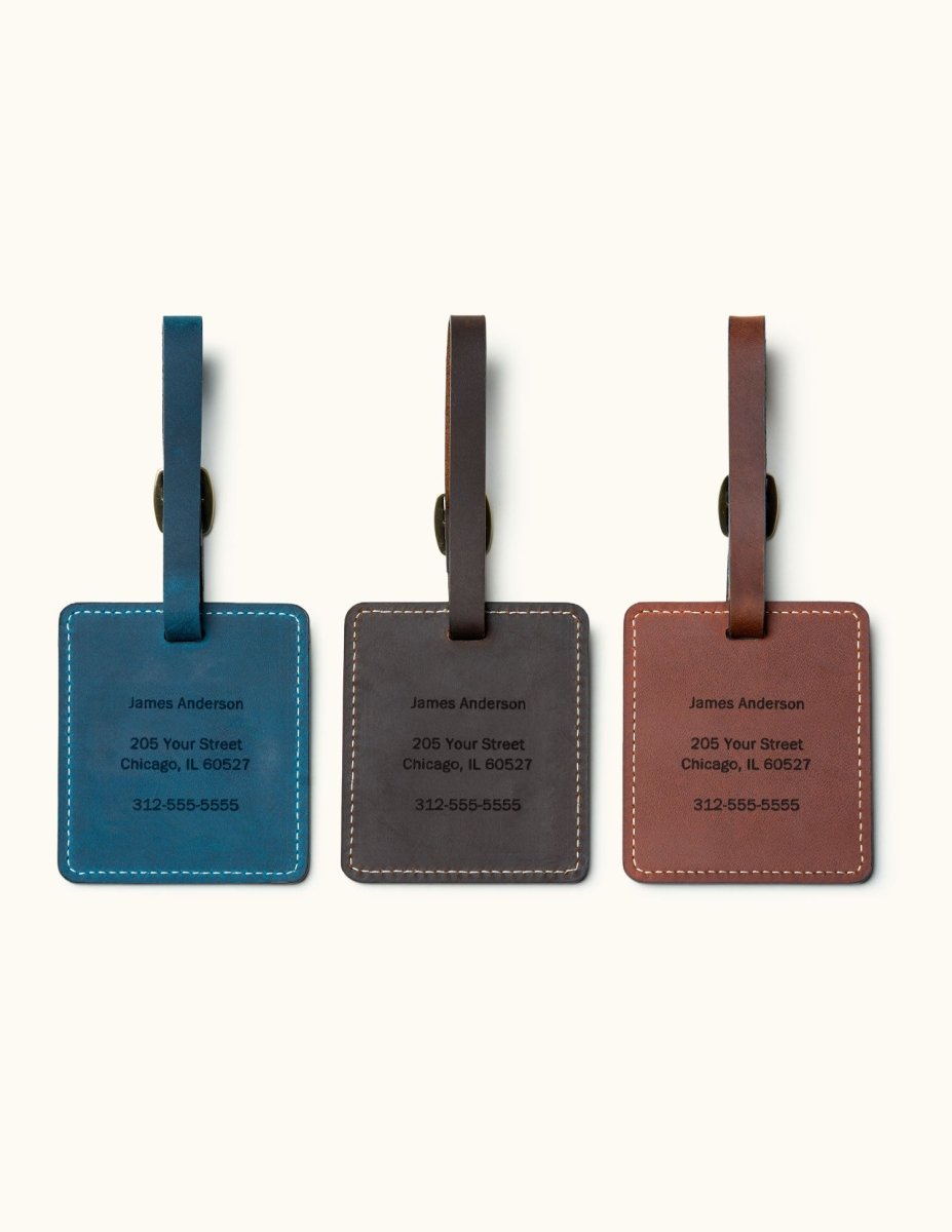 Three handcrafted square leather luggage tags in blue, coffee, and brown with custom engraved personal contact information, white stitching, and brass buckle straps for travel bag identification.