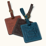 Three handmade square leather luggage tags in vintage brown, coffee, and navy with embossed travel quotes and brass buckles, perfect for identifying travel bags.