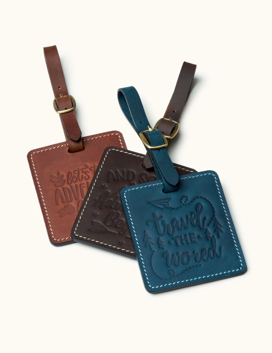 Three handmade square leather luggage tags in vintage brown, coffee, and navy with embossed travel quotes and brass buckles, perfect for identifying travel bags.
